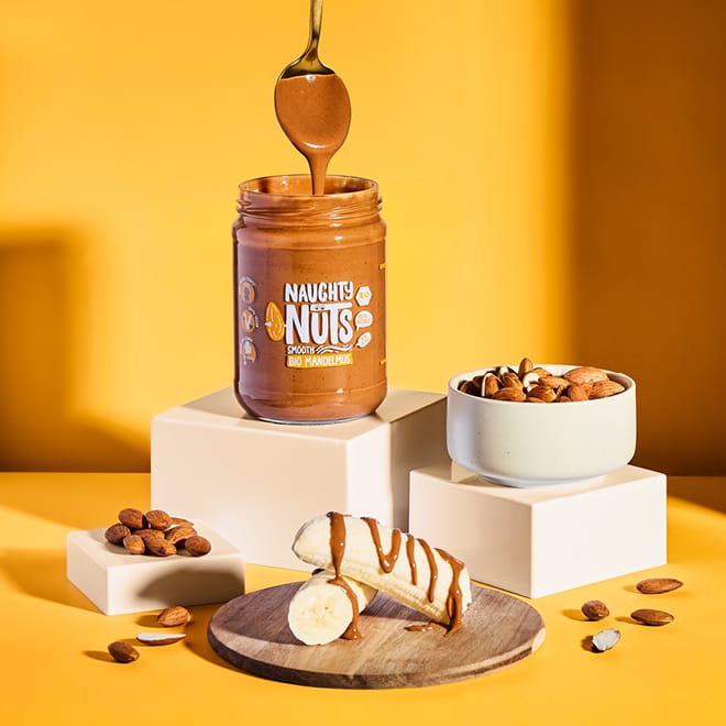 Löffel taucht in ein Glas Bio Mandelmus Smooth, daneben Banane mit Mus und Mandeln