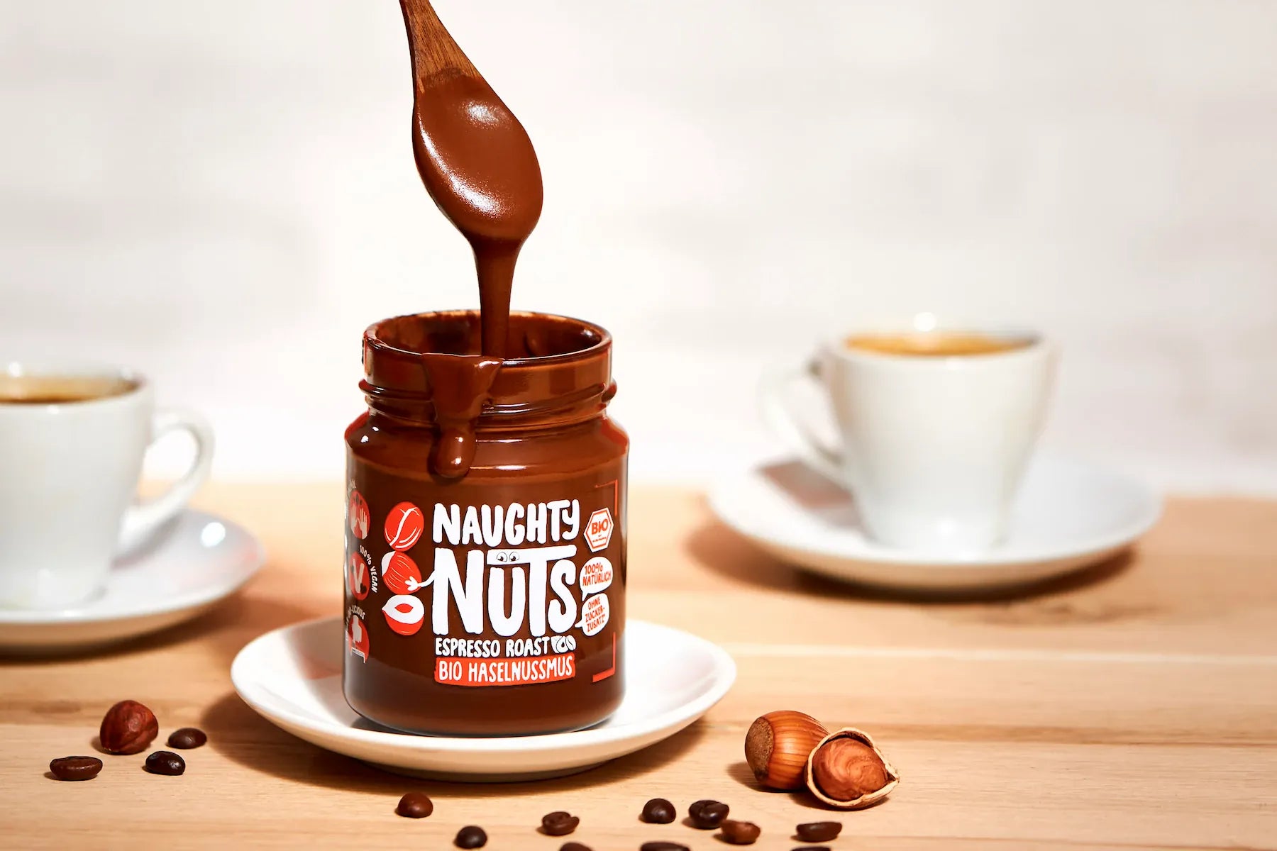 Naughty Nuts Espresso Röstung Bio Haselnussmus im Glas mit Löffel, Kaffeebohnen, Haselnüssen und zwei Tassen Kaffee